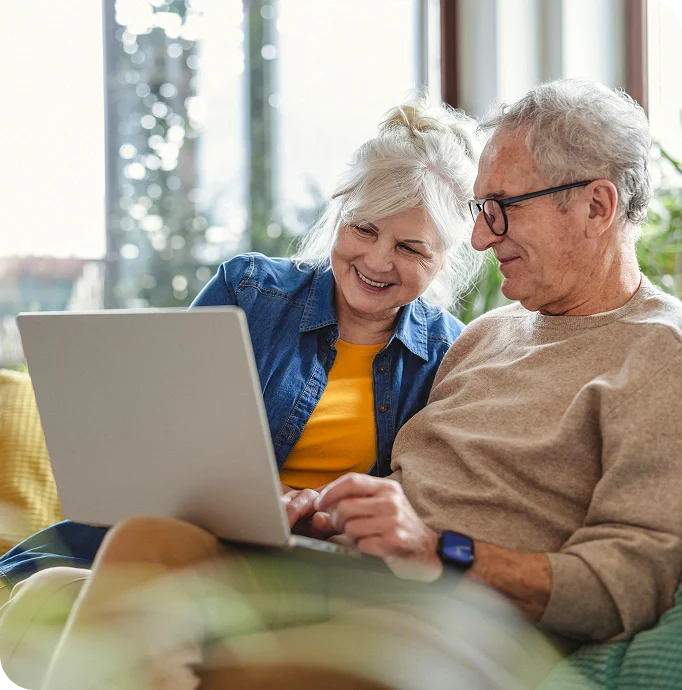 A woman and man sit closely together on a couch, using a laptop in a bright, cozy indoor setting