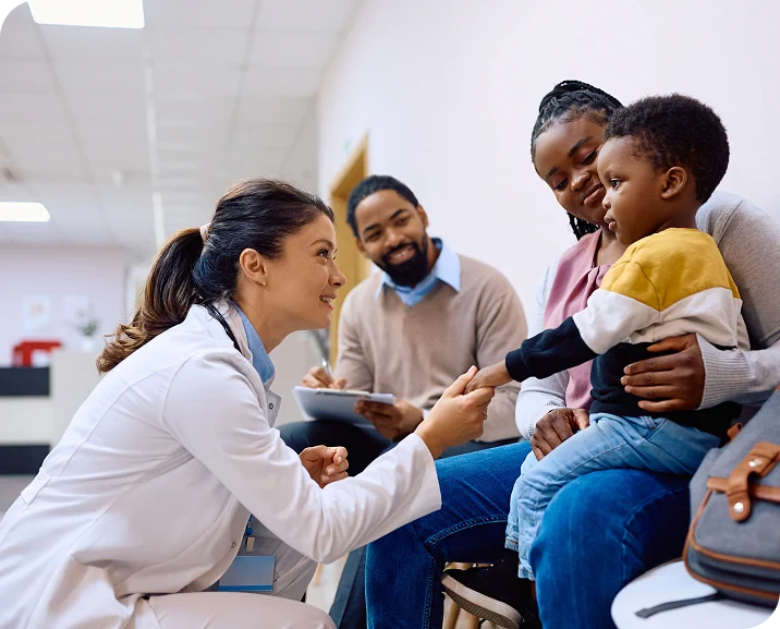 A doctor interacts with a young child while family members observe in a waiting area of a healthcare facility